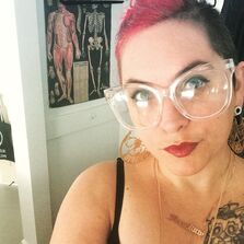 White non-binary femme person with hot pink hair and clear framed glasses. Smirking in front of an anatomy photo while wearing gold earrings.